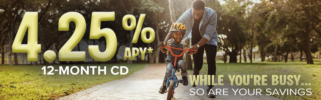 12 MO CD - Community West Credit Union Father helping his young son ride a bike in a sunny park. Ad promotes a 12-month CD at 4.25% APY with the message, “While you’re busy, so are your savings.”