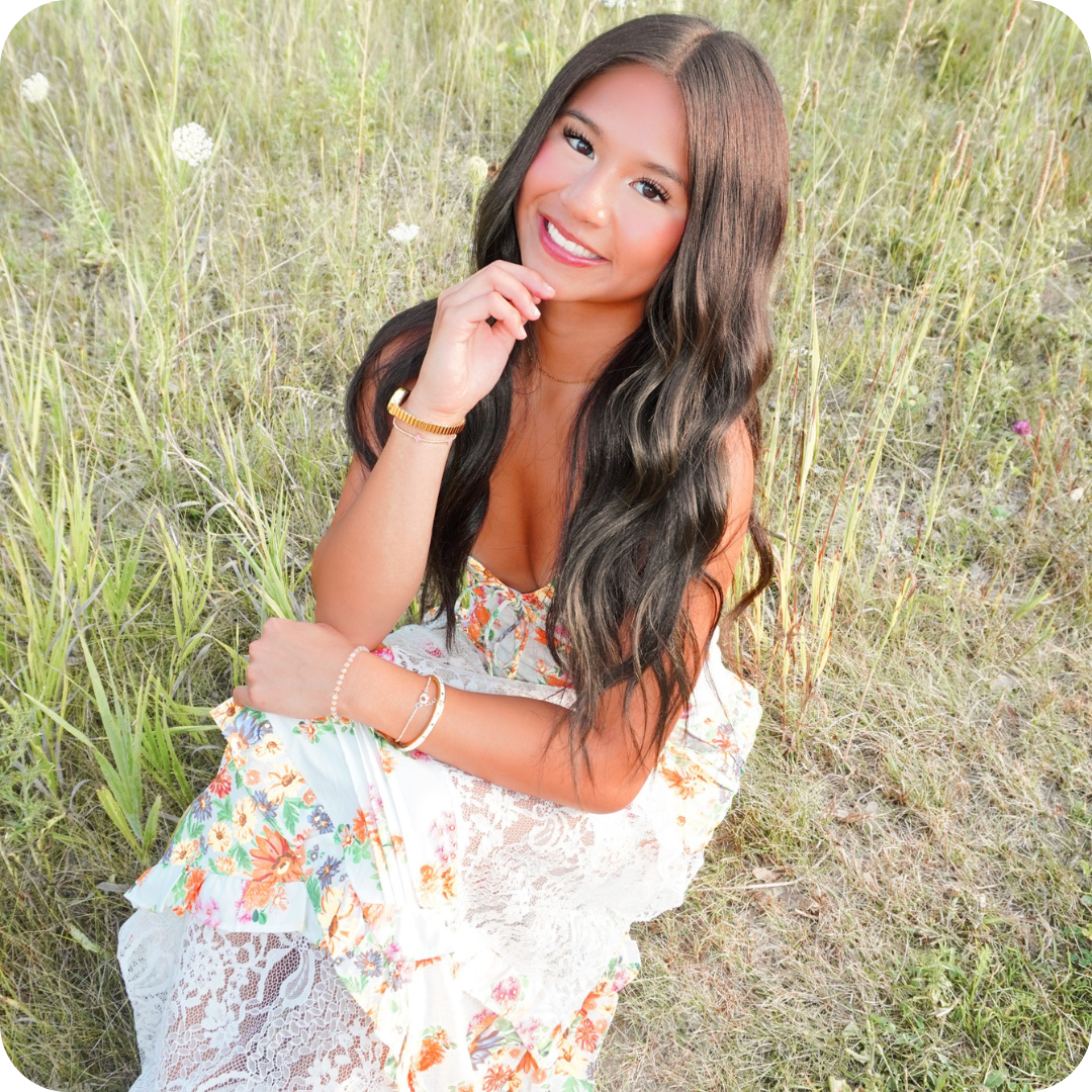 Smiling young woman with long dark hair in a floral dress sitting in a grassy field with wildflowers