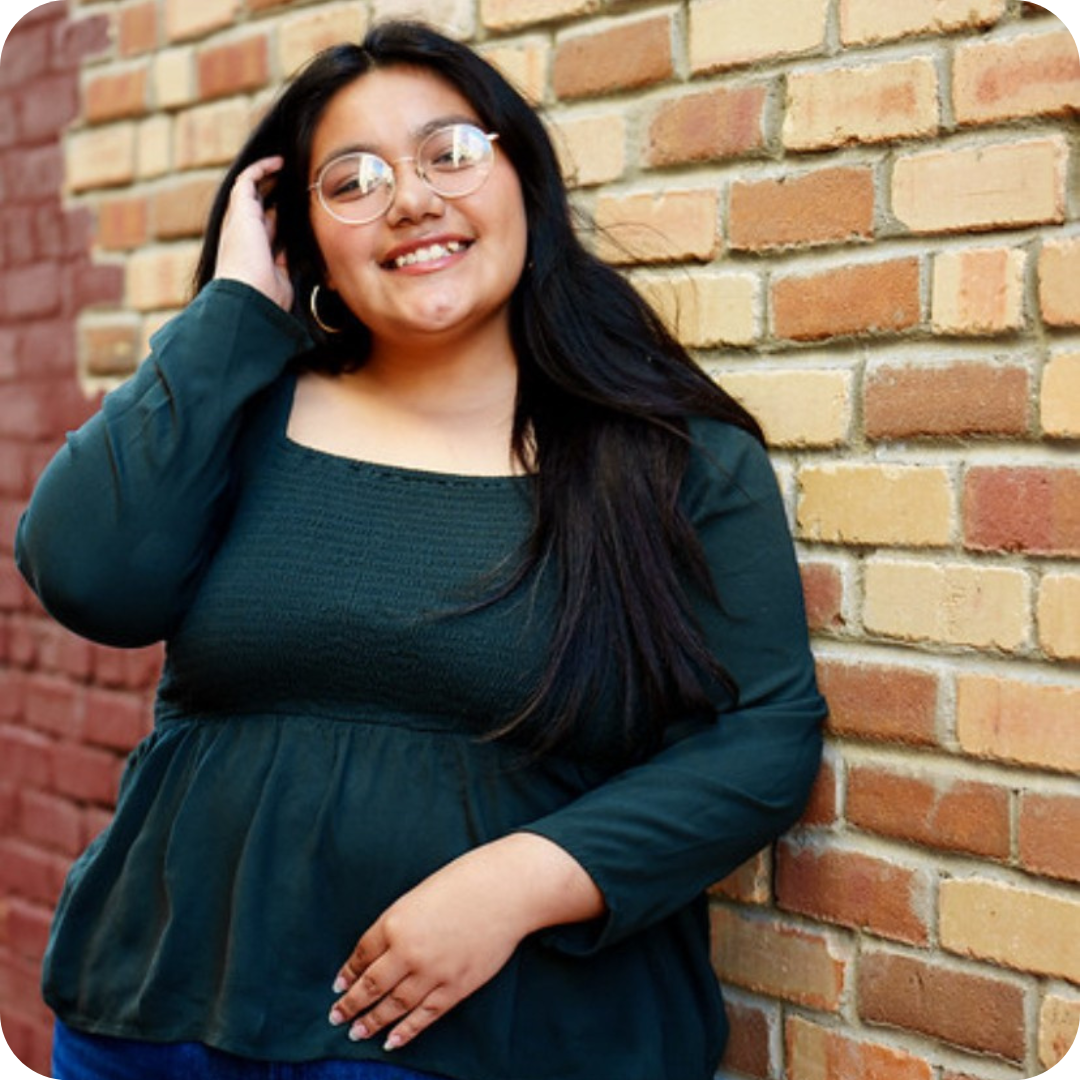 Smiling woman with long dark hair and round glasses standing against a brick wall.