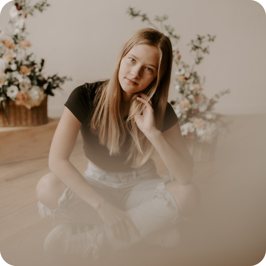 Cecilia-Legacy - Community West Credit Union Young woman with long blond hair sits on a wooden floor, black tee and ripped jeans, soft floral arrangements in the background.