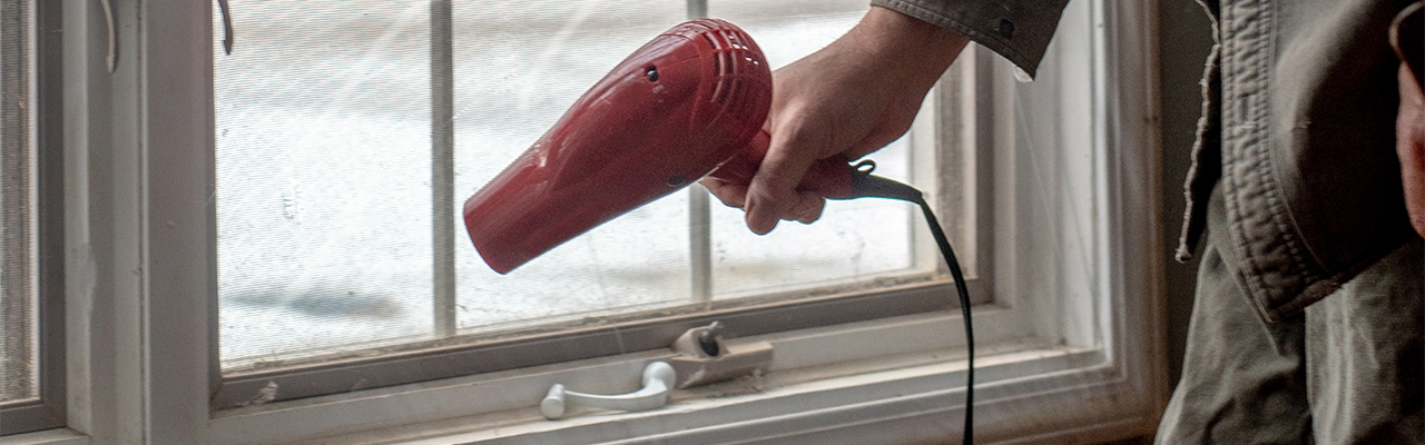 A hand holds a blow dryer and is being used to Shrink wrap a window, to help insulate and keep in the heat for winter